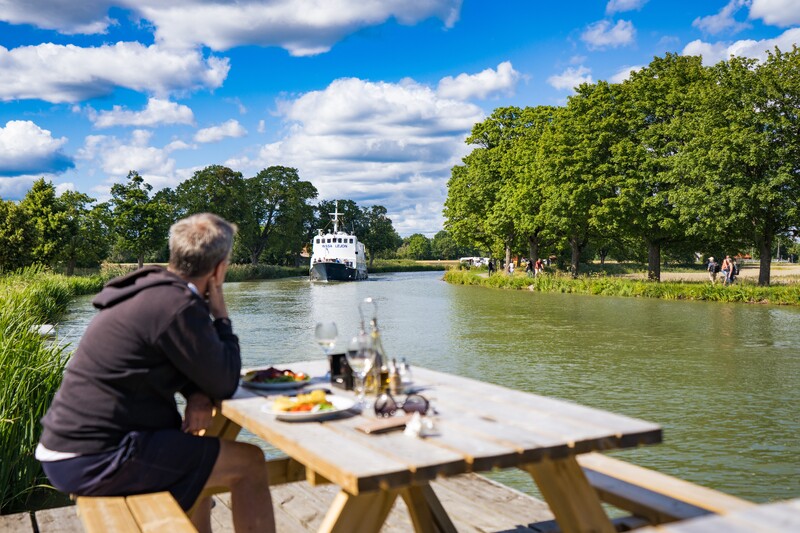 En man äter lunch bredvid kanalen och tittar på en båt som åker förbi